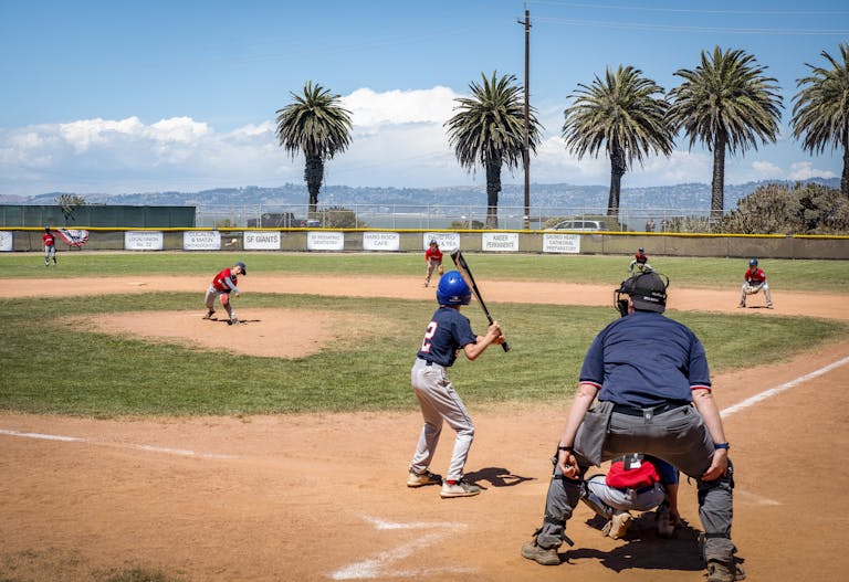 Children playing baseball in an outdoor field under a bright blue sky with palm trees in the background.